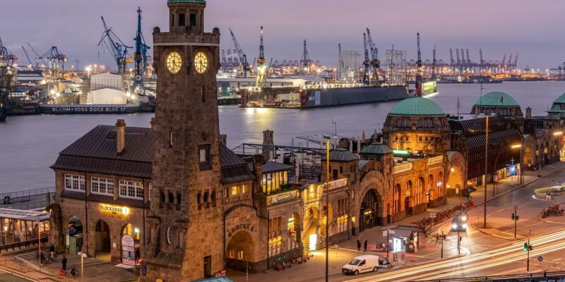 Ein ruhiger Abendblick auf die St. Pauli Landungsbrücken in Hamburg, Deutschland. Der Uhrenturm ist auffällig beleuchtet, die umliegenden Gebäude sind ebenfalls beleuchtet. Vor dem Abendhimmel bilden der Hamburger Hafen und die Kräne eine beeindruckende Kulisse, während Autos und Straßenlaternen die Straßen säumen.