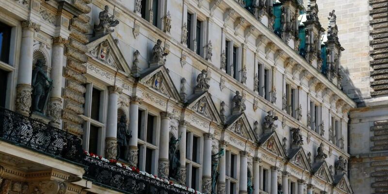 Das Hamburger Rathaus besticht durch eine detailreiche Fassade mit Reihen verzierter Fenster und Statuen in Nischen im klassischen Stil. Seine Architektur umfasst aufwendige Steinmetzarbeiten und ein dekoratives schwarzes Geländer, das mit kleinen Pflanzen geschmückt ist und die Erhabenheit dieses historischen Gebäudes widerspiegelt.