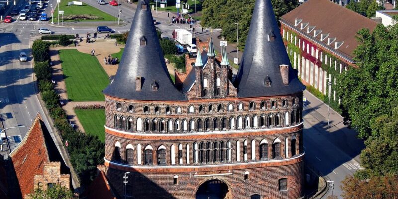 Luftaufnahme des Holstentors in Lübeck, einem historischen Backsteintor mit zwei runden Türmen und einem gewölbten Eingang. Es ist umgeben von belebten Straßen, sattgrünen Rasenflächen und nahe gelegenen Gebäuden, die den Charme Lübecks widerspiegeln.