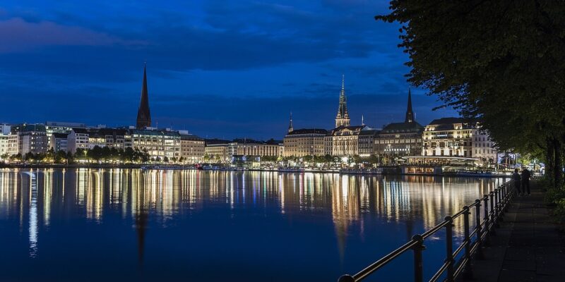 Eine nächtliche Stadtansicht von Hamburg, Deutschland, zeigt die beleuchteten Gebäude und Kirchtürme des Jungfernstiegs, die sich im ruhigen Wasser der Alster spiegeln. Der Himmel ist tiefblau, und auf der rechten Seite des Bildes säumen Bäume das Bild.