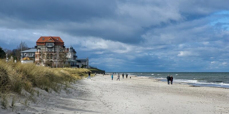Ein Sandstrand am Kühlungsborner Strand mit ein paar Leuten, die unter einem bewölkten Himmel am Ufer entlangspazieren. Links steht ein großes, reich verziertes Gebäude mit roten Dächern nahe dem Rand des Strandes, umgeben von grasbewachsenen Dünen. Das Meer ist ruhig mit sanften Wellen.
