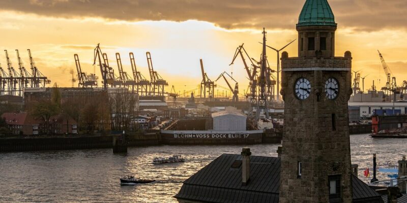 Eine Szene am Flussufer bei Sonnenuntergang mit dem ikonischen Uhrturm der Landungsbrücken im Vordergrund. Im Hintergrund sind mehrere Kräne auf einer Werft unter einem warmen, leuchtenden Himmel zu sehen. Das Wasser spiegelt die goldenen Töne des Sonnenuntergangs wunderschön wider.