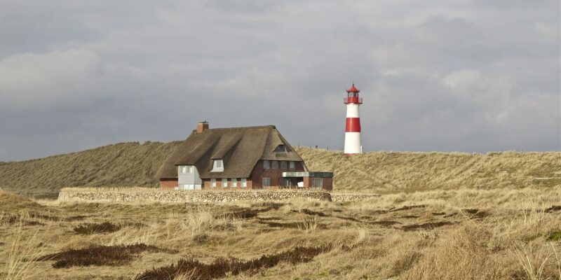 Auf einer Wiese steht ein reetgedecktes Haus. In der Ferne ist der Leuchtturm Lister Ellenbogen auf Sylt zu sehen, dessen rote und weiße Streifen einen Kontrast zum teilweise bewölkten Himmel bilden.