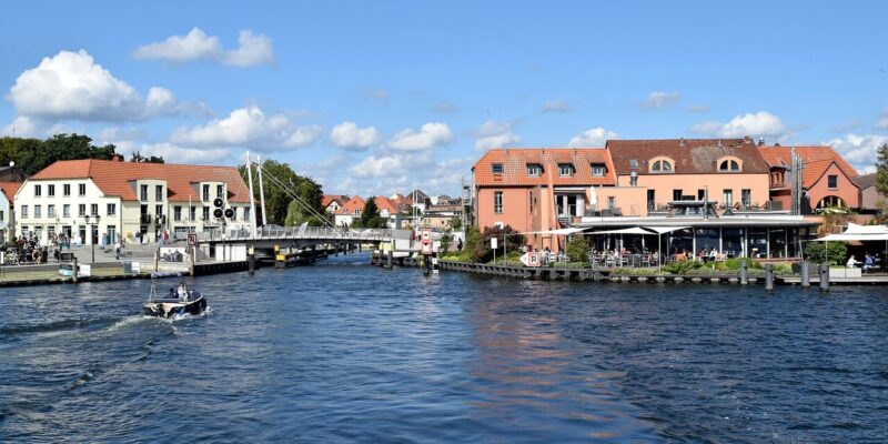 Ein malerischer Blick auf den Fluss in Malchow mit einem kleinen Boot, das sanft durch das Wasser gleitet. Auf der rechten Seite säumen charmante Gebäude mit Sitzgelegenheiten im Freien die Uferpromenade, während sich eine malerische Brücke anmutig unter einem klaren blauen Himmel mit vereinzelten Wolken wölbt.