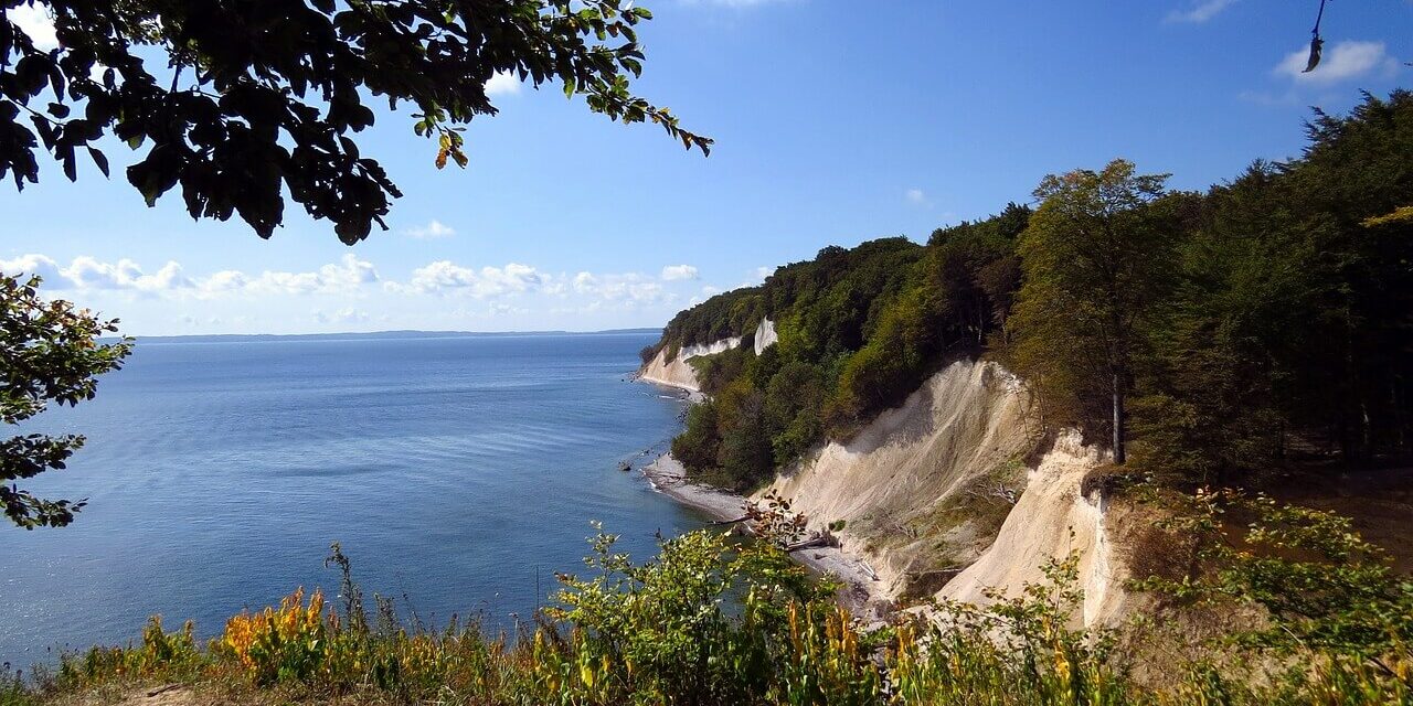 Mecklenburg-Vorpommern Nationalpark Malerischer Blick auf weiße Kreidefelsen in einem der atemberaubenden Nationalparks in Mecklenburg-Vorpommern. Üppiges Grün bedeckt die Klippen unter einem strahlend blauen Himmel mit Wolken, während sich das ruhige Meer bis zum Horizont erstreckt. Im Vordergrund sind grüne Blätter und ein Fleck mit bunten Wildblumen zu sehen.