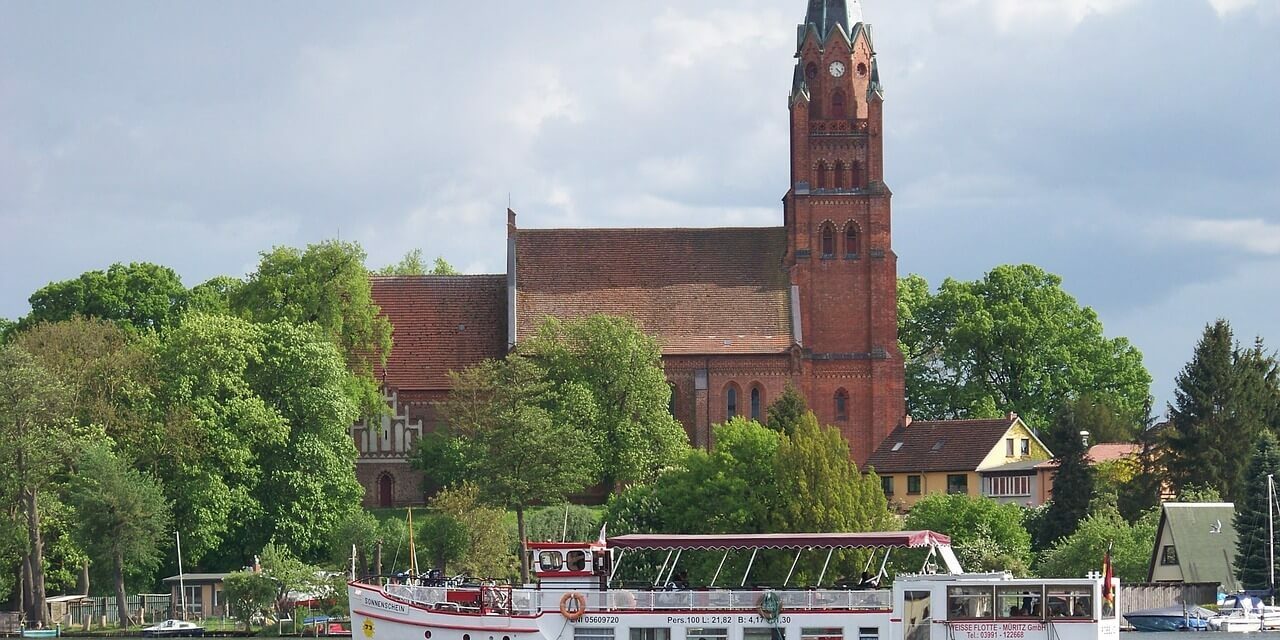 Ein weißes Ausflugsboot mit Passagieren fährt auf einem Fluss in der Mecklenburgischen Seenplatte und passiert eine historische rote Backsteinkirche mit einem hohen Kirchturm. Die Kirche ist elegant von üppigen grünen Bäumen unter einem wolkigen Himmel eingerahmt.
