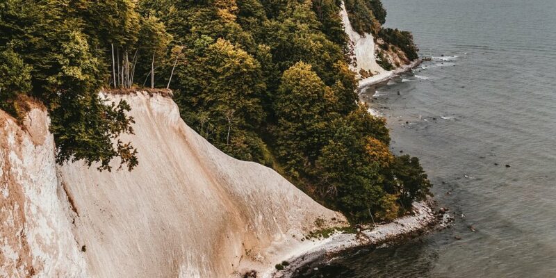 Ein malerischer Blick auf weiße Kreidefelsen entlang der Küste des Nationalparks Jasmund, gesäumt von dichten grünen Wäldern. Die Klippen überblicken ein ruhiges, ausgedehntes Gewässer, wobei sich die Uferlinie unter einem bewölkten Himmel in die Ferne wölbt.