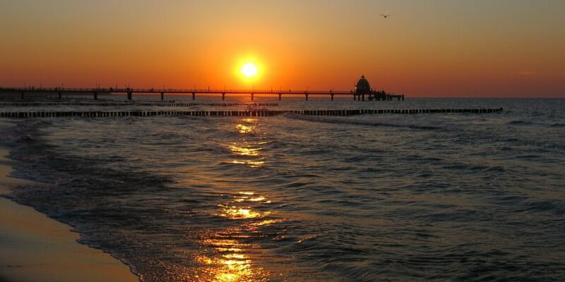 Ein ruhiger Sonnenuntergang über dem Ostseestrand Zingst. Die tief am Himmel stehende Sonne wirft einen goldenen Widerschein auf das Wasser. Ein Pier ragt ins Meer und zeichnet sich als Silhouette gegen die warmen Farben des Himmels ab, während sanfte Wellen an die Küste plätschern.