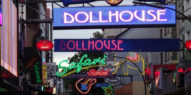 Eine belebte Straße mit zahlreichen leuchtenden Neonschildern, darunter Dollhouse und Safari, erhellt die Nacht auf der Reeperbahn in Hamburg. Diese lebendige Szene fängt die farbenfrohe Atmosphäre eines urbanen Nachtlebenviertels voller Energie und Aufregung ein.
