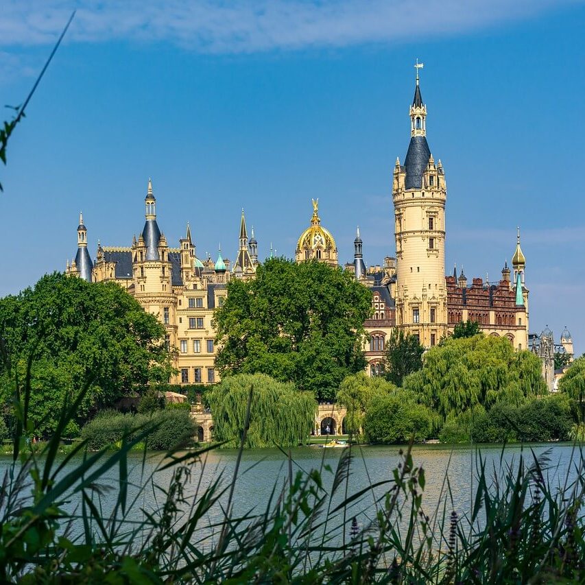 Ein malerischer Blick auf Schloss Schwerin mit seinen verzierten Türmen und Spitzen vor einem klaren blauen Himmel. Im Vordergrund sind üppiges Grün und ein ruhiger See zu sehen, was das märchenhafte Aussehen des Schlosses noch verstärkt und es zu einem der bezauberndsten Schlösser in Mecklenburg-Vorpommern macht.