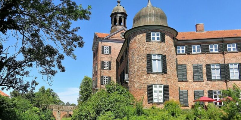 Das rote Backsteingebäude von Schloss Eutin mit seiner Kuppel und dem Glockenturm thront über üppigen grünen Gärten neben einem Kanal. Eine Steinbogenbrücke überquert das Wasser und der klare blaue Himmel unterstreicht diese ruhige Umgebung.
