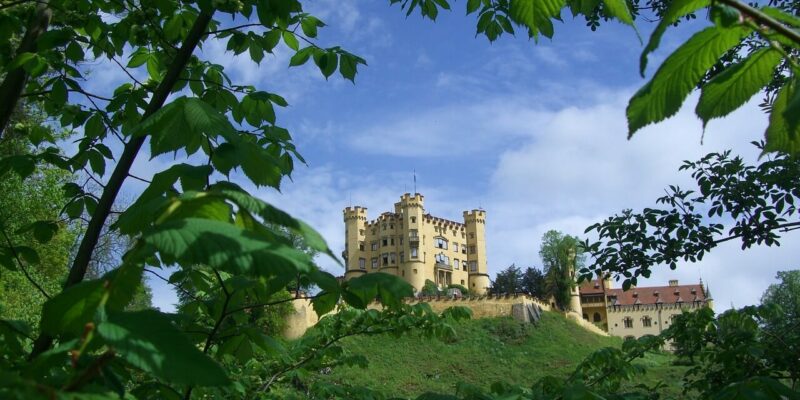 Auf einem Hügel steht ein gelbes Schloss mit mehreren Türmen, teilweise verdeckt von üppigen grünen Blättern. Das Schloss Hohenschwangau ist an einem hellen Tag unter einem mit weißen Wolken übersäten Himmel zu sehen.