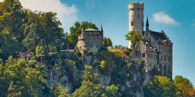 Schloss Lichtenstein liegt auf einem felsigen, bewaldeten Hügel unter einem strahlend blauen Himmel und ist eine mittelalterliche Burg mit einem runden Turm und steilen Dächern. Üppige grüne Bäume umgeben das historische Bauwerk und bilden eine malerische und ruhige Landschaft.