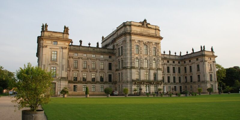 Ein großes, historisches Steingebäude mit kunstvollen architektonischen Details steht vor einem klaren Himmel. Das Gebäude, das an Schloss Ludwigslust erinnert, weist Statuen entlang der Dachlinie auf. Im Vordergrund befindet sich ein gepflegter Rasen mit Topfbäumen.