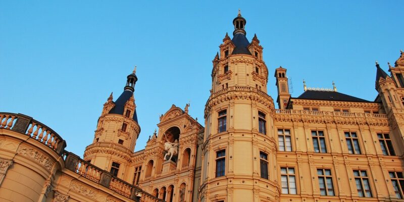 Ein majestätisches, reich verziertes Schloss, das an Schloss Schwerin erinnert, steht mit hohen, spitzen Türmen und komplizierten architektonischen Details vor einem klaren blauen Himmel. Seine Struktur zeichnet sich durch kunstvolle Fenster und dekorative Elemente in einer warmen, beigen Steinfassade aus.