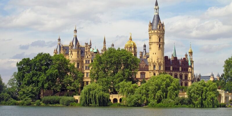 Das prächtige Schweriner Schloss mit seinen zahlreichen Türmen und der goldenen Kuppel steht majestätisch inmitten üppiger Grünflächen in der Nähe eines ruhigen Sees. Der teilweise bewölkte Himmel verstärkt die ruhige Atmosphäre dieser malerischen Szene.