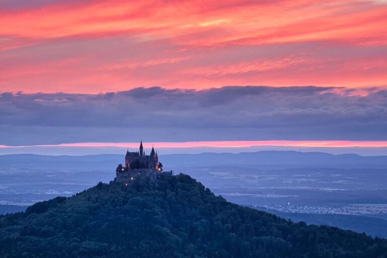 Ein malerisches Schloss, das an die Burg Hohenzollern erinnert, thront auf einem bewaldeten Hügel und hebt sich vor einem dramatischen Sonnenuntergangshimmel mit leuchtenden Rosa- und Orangetönen ab. Im Hintergrund erstrecken sich sanfte Hügel und ferne Landschaften und schaffen eine ruhige und majestätische Szene, die typisch für Sehenswürdigkeiten in Baden-Württemberg ist.
