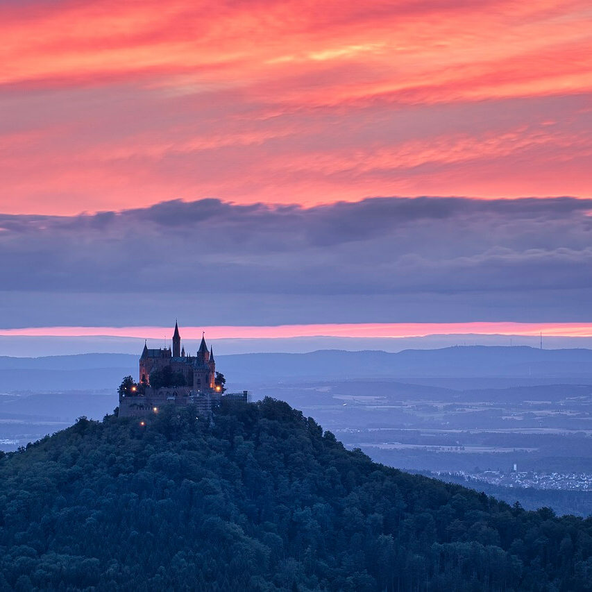 Ein malerisches Schloss, das an die Burg Hohenzollern erinnert, thront auf einem bewaldeten Hügel und hebt sich vor einem dramatischen Sonnenuntergangshimmel mit leuchtenden Rosa- und Orangetönen ab. Im Hintergrund erstrecken sich sanfte Hügel und ferne Landschaften und schaffen eine ruhige und majestätische Szene, die typisch für Sehenswürdigkeiten in Baden-Württemberg ist.