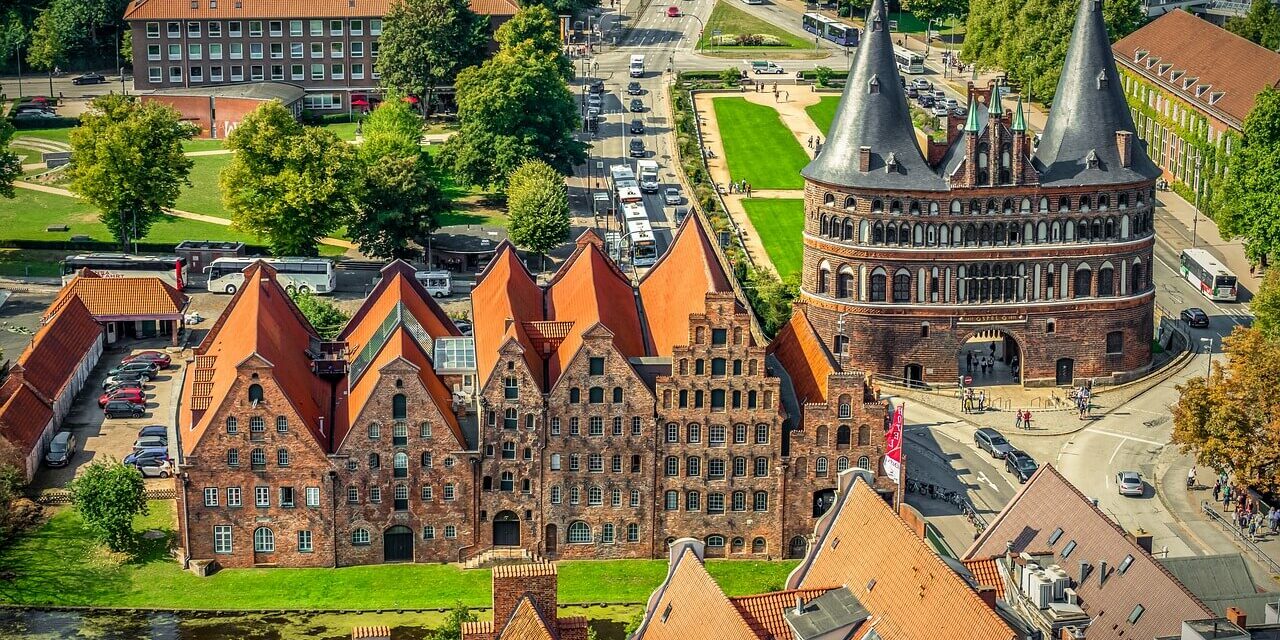 Luftaufnahme von Lübeck, einem Highlight unter den Sehenswürdigkeiten in Schleswig-Holstein, mit mittelalterlicher Architektur mit roten Backsteingebäuden und dem ikonischen Holstentor. Die alten Lagerhäuser und grünen Rasenflächen sind von Straßen und Bäumen unter einem klaren Himmel umgeben.