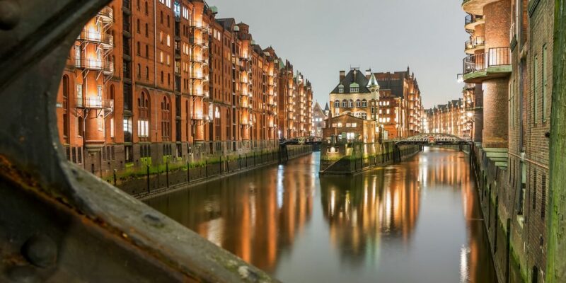 Eine nächtliche Szene der Speicherstadt, einem historischen Viertel mit beleuchteten roten Backsteingebäuden, die einen ruhigen Kanal säumen und das warme Licht reflektieren. In der Mitte des Kanals steht ein Gebäude, das von einer Metallbrücke aus sichtbar ist.