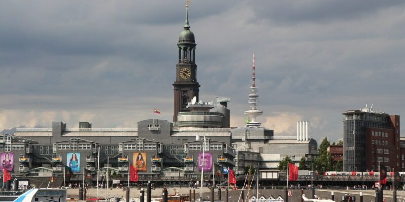 Eine Stadtansicht mit einer Uferpromenade und angedockten Booten. Im Hintergrund erhebt sich die St. Michaelis Kirche in Hamburg über modernen Gebäuden. Flaggen schmücken die Gegend und der Himmel ist mit Wolken bedeckt.