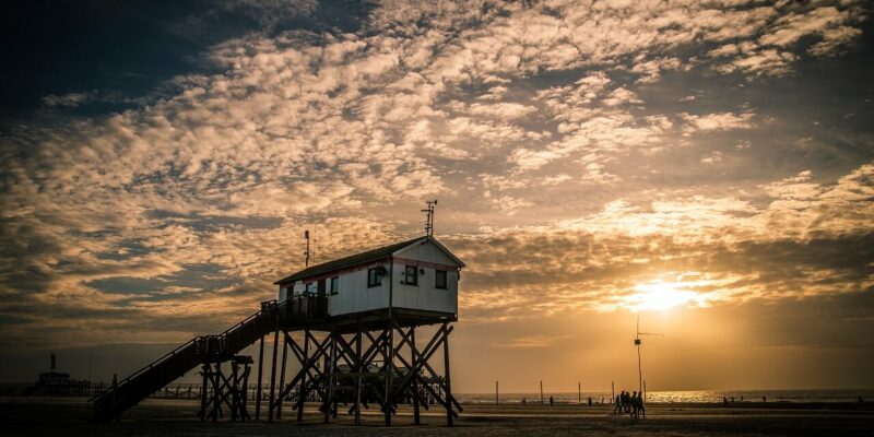 Eine hölzerne Rettungsschwimmerstation auf Stelzen steht bei Sonnenuntergang am Sandstrand von St. Peter Ording. Der Himmel ist voller dramatischer Wolken und die Sonne lugt nahe dem Horizont hervor und wirft einen warmen Schein auf die Szene mit dem Meer im Hintergrund.