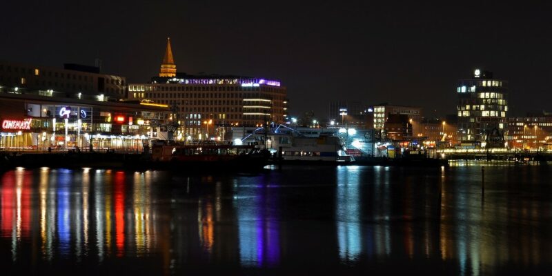 Eine nächtliche Stadtansicht der Stadt Kiel zeigt Gebäude mit verschiedenen Lichtern, die sich auf einer ruhigen Wasseroberfläche spiegeln. Neonschilder und beleuchtete Fenster erzeugen farbenfrohe Reflexionen, während in der Ferne ein hohes, spitzes Gebäude sichtbar ist.