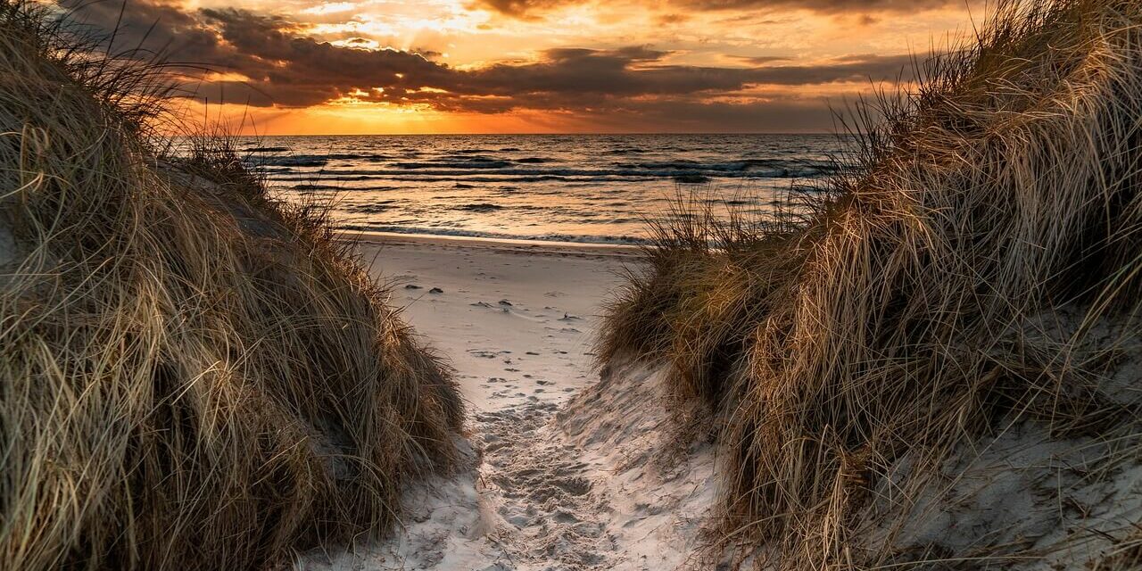 Ein schmaler Sandweg schlängelt sich durch grasbewachsene Dünen zu einem Strand im Sonnenuntergang, der an die ruhigen Strände in Mecklenburg-Vorpommern erinnert. Die Sonne geht über dem Meer unter und wirft warme orangefarbene und goldene Farbtöne in den Himmel und auf die Wolken, während die Wellen sanft das Ufer berühren.