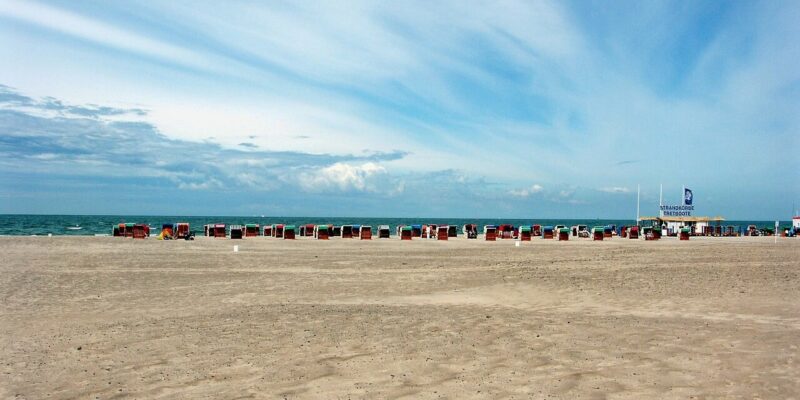 Am Strand von Warnemünde liegt ein breiter Sandstrand mit verstreuten Strandkörben am Meer. Der Himmel ist teilweise bewölkt und weist blaue und weiße Streifen auf. In der Ferne, wo das Meer unter einem weiten Himmel auf den Horizont trifft, erwartet Sie Ruhe.