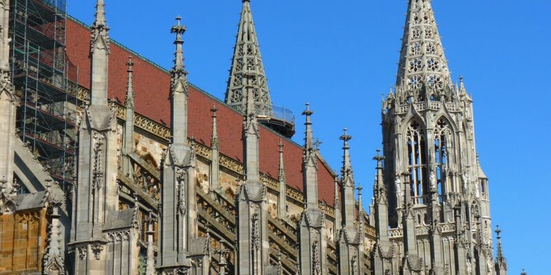 Das Ulmer Münster, eine beeindruckende Kathedrale im gotischen Stil mit kunstvollen Türmen und detailreichem Mauerwerk, steht majestätisch unter einem klaren blauen Himmel. Auf einer Seite sind Gerüste sichtbar, die auf laufende Restaurierungsarbeiten hinweisen, um die historische Pracht zu bewahren.