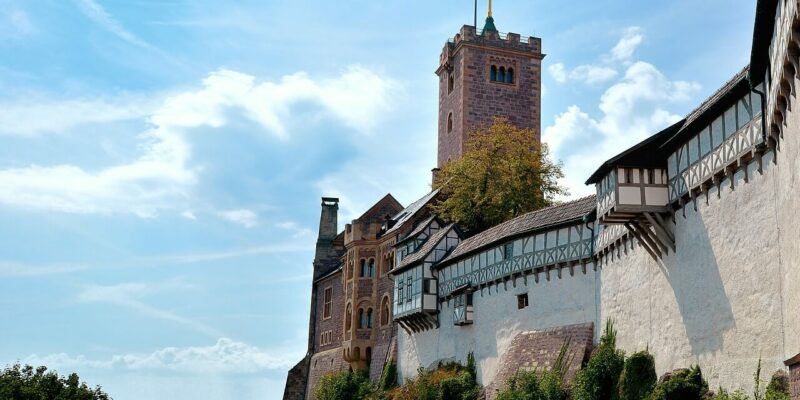 Die Wartburg liegt auf einem Hügel und ist eine historische Steinburg mit Fachwerkmauern und einem markanten Turm, auf dem eine Flagge weht. Sie ragt stolz vor einem strahlend blauen Himmel mit flauschigen Wolken empor und ist von üppigem Grün umgeben.